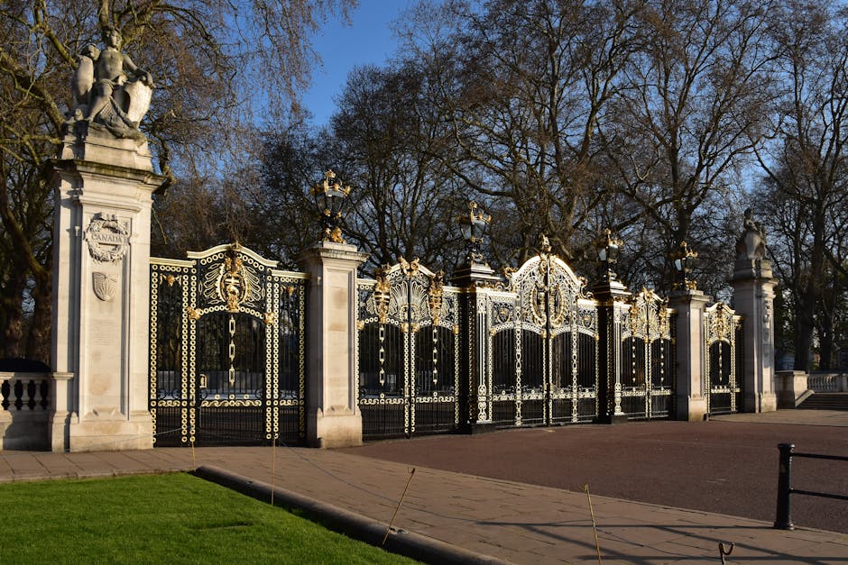 An ornate wrought iron gate with intricate gold and black detailing, supported by tall, white stone pillars topped with classical statues, is part of a decorative entrance to a park or estate. Behind