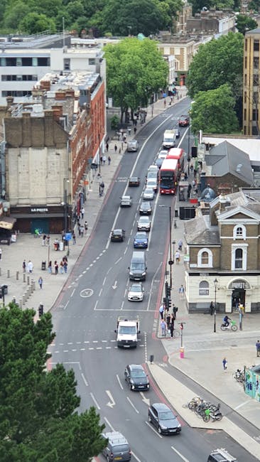 An aerial view of a busy urban street scene with parked and moving cars, including a red double-decker bus and several smaller vehicles, along with pedestrians walking on the sidewalks and crossing at