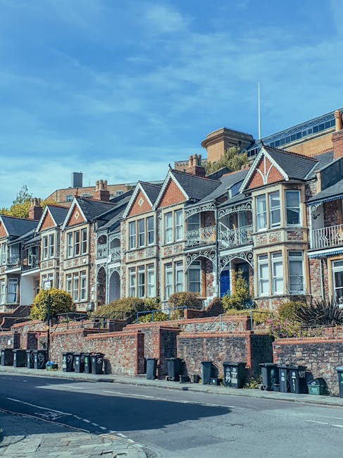 A row of historic terraced houses with ornate Victorian-style architecture, featuring decorative ironwork balconies, bay windows, and gabled roofs, situated along a street with a brick retaining wall